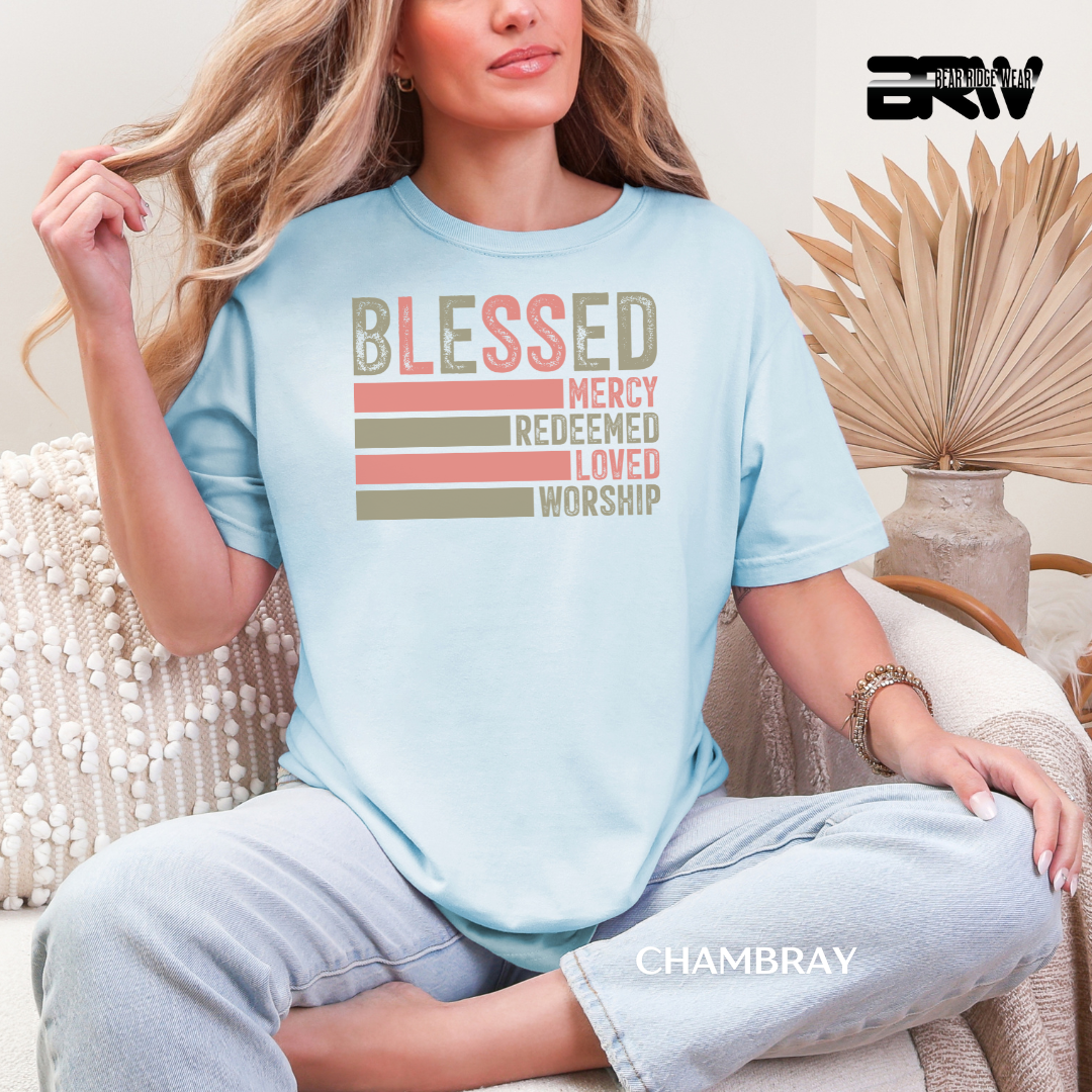 Woman wearing a light blue t-shirt with motivational text, sitting in a cozy room. BLESSED, MERCY REDEEMED, LOVED AND WORSHIP FAITH TEE