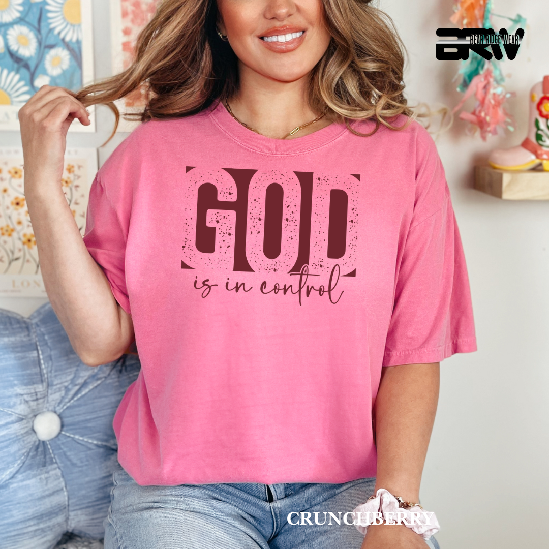 Woman wearing a pink t-shirt with 'GOD is in control' text, sitting on a couch.