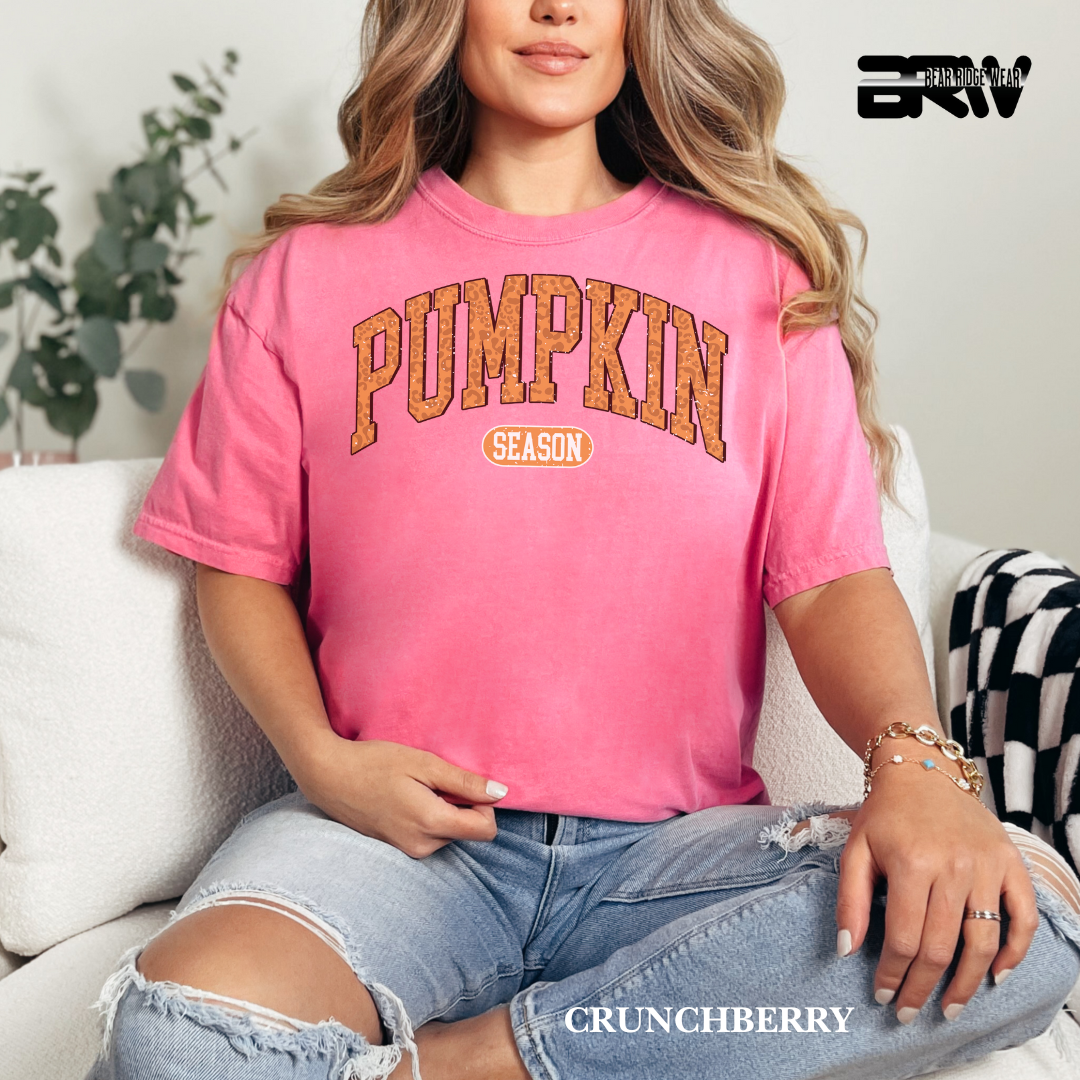 Woman wearing a pink 'Pumpkin Season' t-shirt sitting on a couch with a plant in the background.