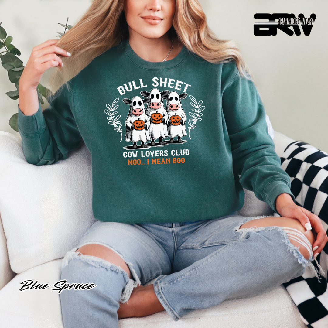 Person wearing a green sweatshirt with cow graphics and text, sitting on a couch. FALL AND PUMPKIN TEE