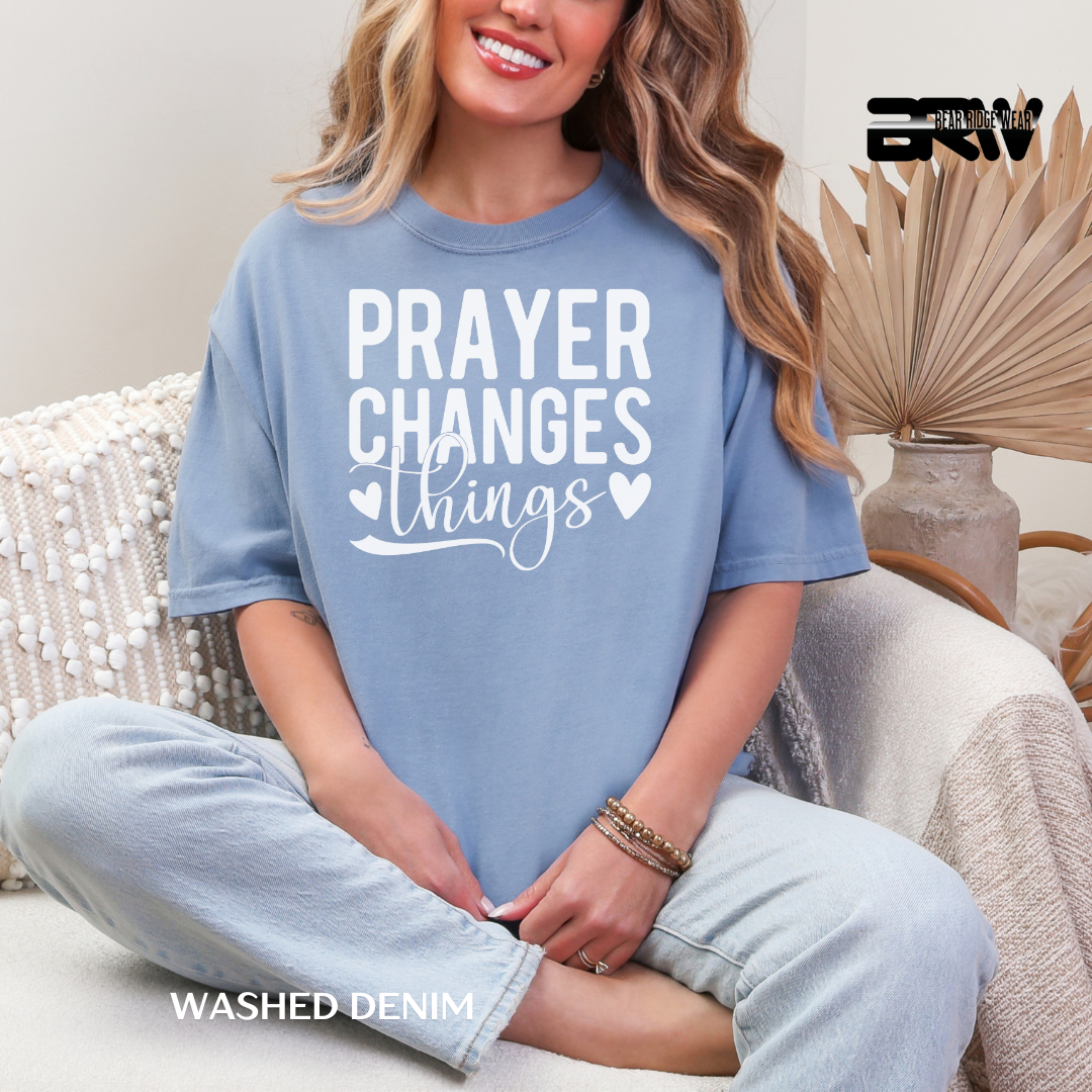 Woman wearing a blue t-shirt with 'PRAYER CHANGES THINGS' text, sitting on a couch.