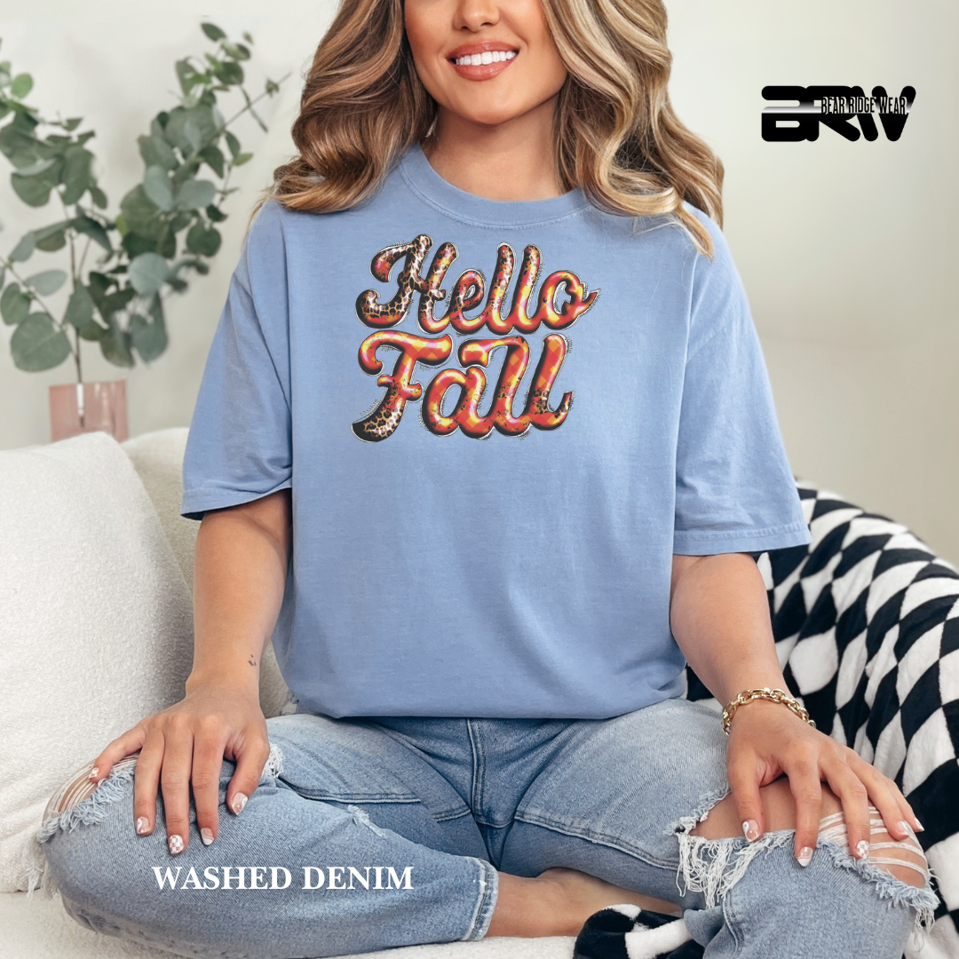 Woman wearing a blue 'Hello Fall' t-shirt sitting on a couch with a plant in the background