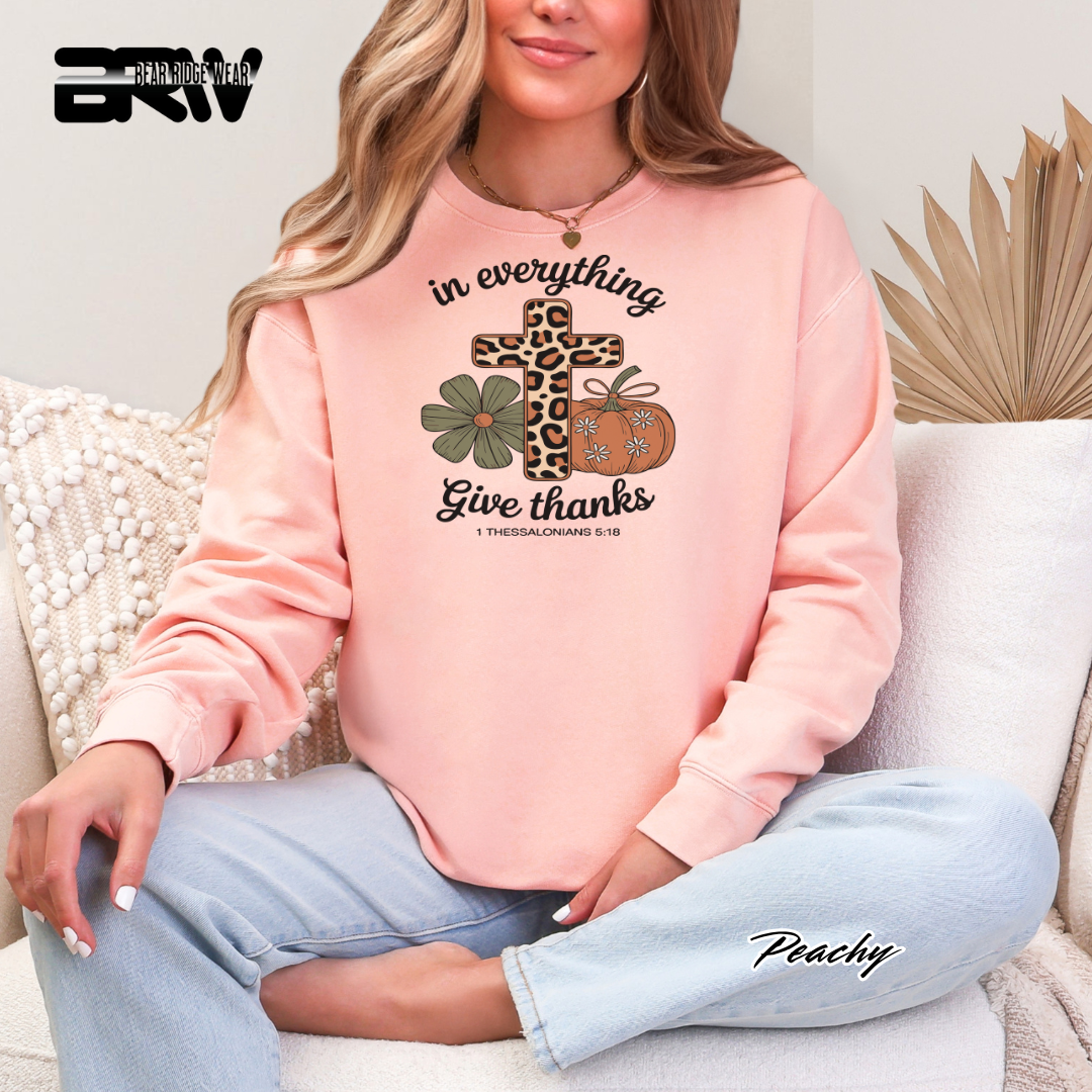 Person wearing a pink sweatshirt with a graphic design and text, sitting on a couch. IN EVERYTHING GIVE THANKS PUMPKIN FALL TEE