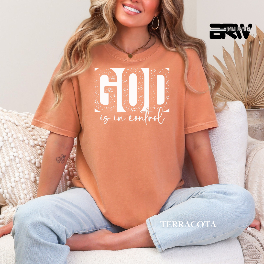 Person wearing a terracotta t-shirt with 'God is in control' text, sitting on a couch.