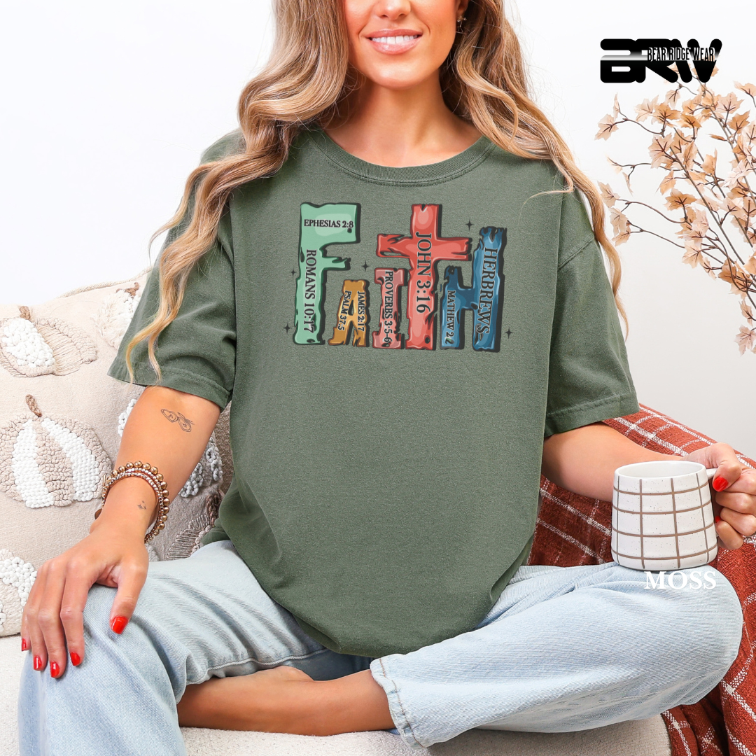 Woman wearing a green t-shirt with colorful text design, sitting on a couch holding a mug. COLORFUL FAITH TEE