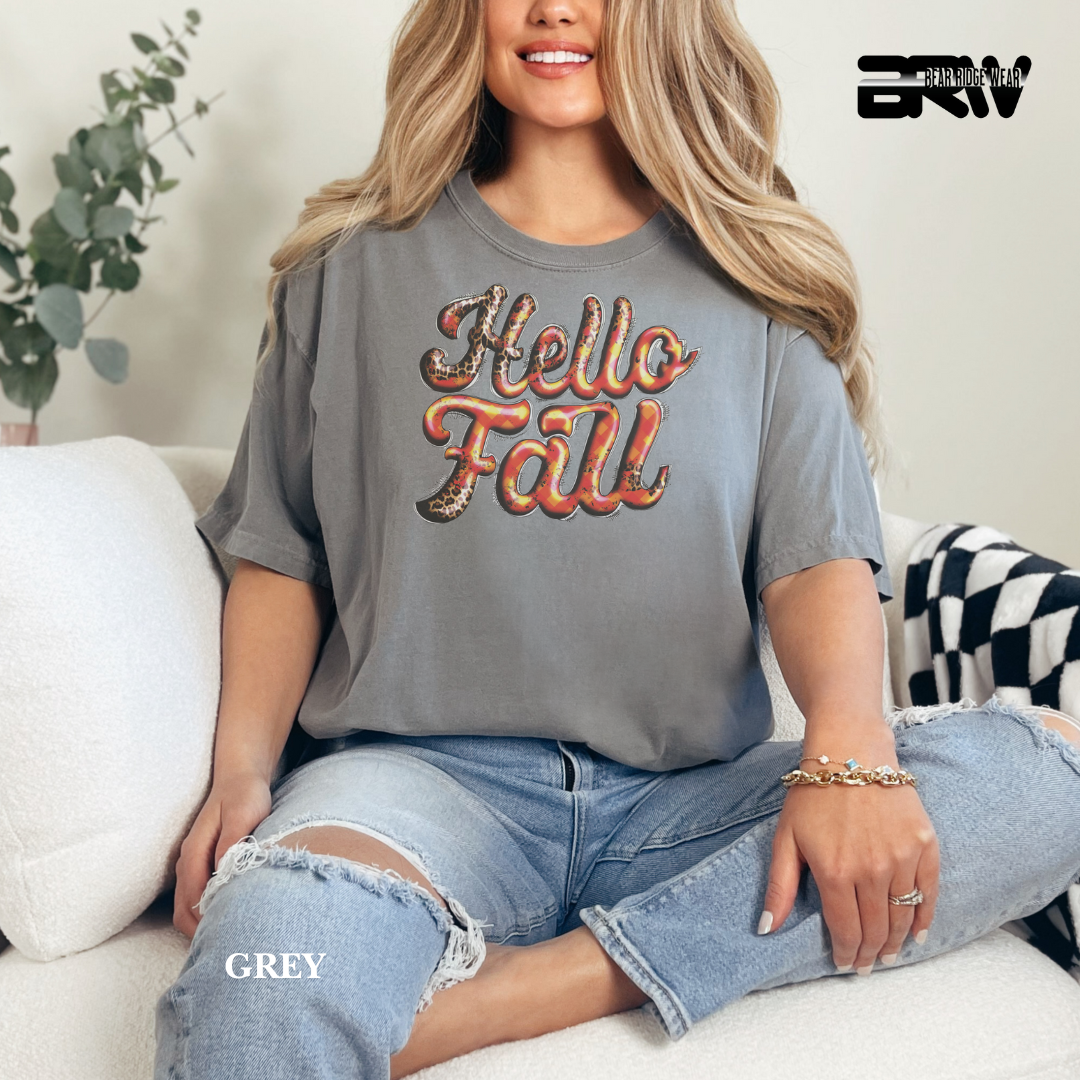 Woman wearing a 'Hello Fall' shirt sitting on a couch with a plant in the background