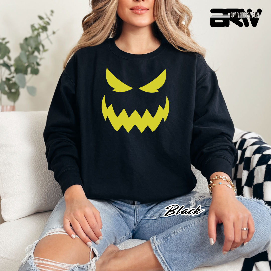 Person wearing a black sweatshirt with a yellow graphic design, sitting on a couch. SCARY PUMPKIN FACE FALL TEE