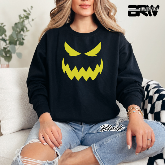 Person wearing a black sweatshirt with a yellow graphic design, sitting on a couch. SCARY PUMPKIN FACE FALL TEE