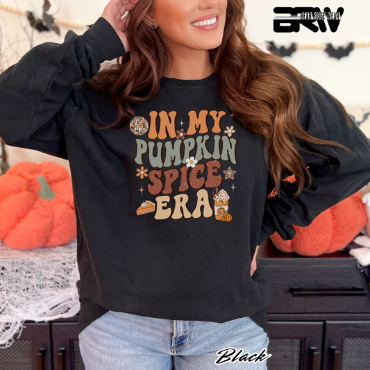 Person wearing a black sweatshirt with 'In My Pumpkin Spice Era' text, standing in front of pumpkins.