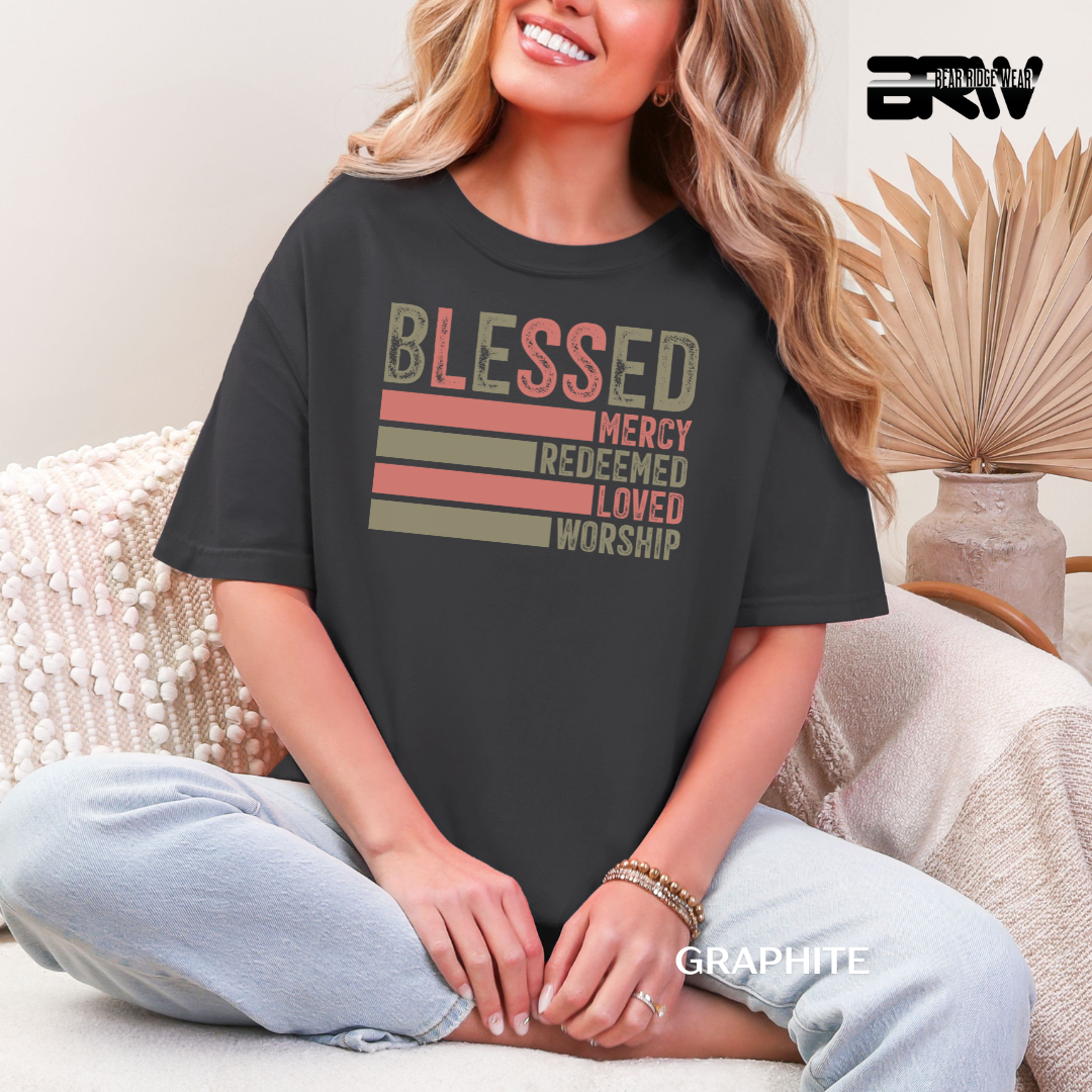Woman wearing a black t-shirt with motivational text, sitting on a couch. BLESSED, MERCY REDEEMED, LOVED AND WORSHIP FAITH TEE