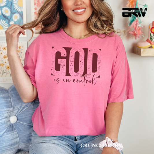 Woman wearing a pink t-shirt with 'GOD is in control' text, sitting on a couch.