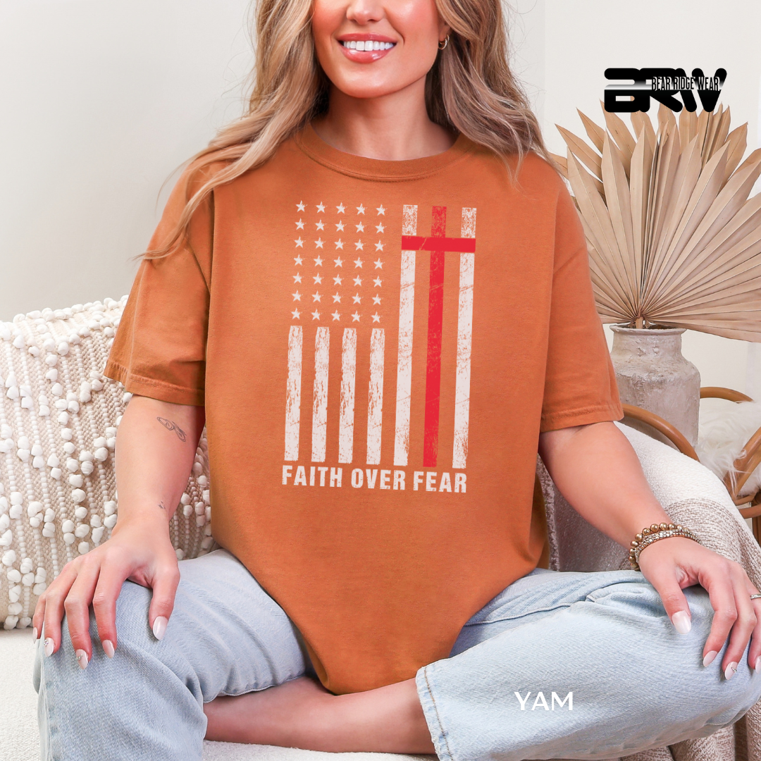 Person wearing an orange t-shirt with a flag design and 'Faith Over Fear' text, sitting on a couch.