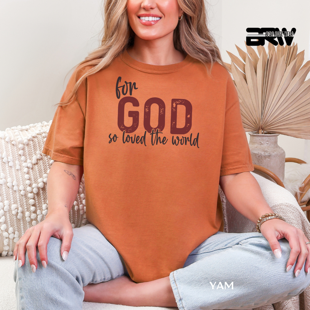 Person wearing a brown t-shirt with text, sitting on a couch in a home setting. FOR GOD SO LOVE THE WORLD FAITH TEE 