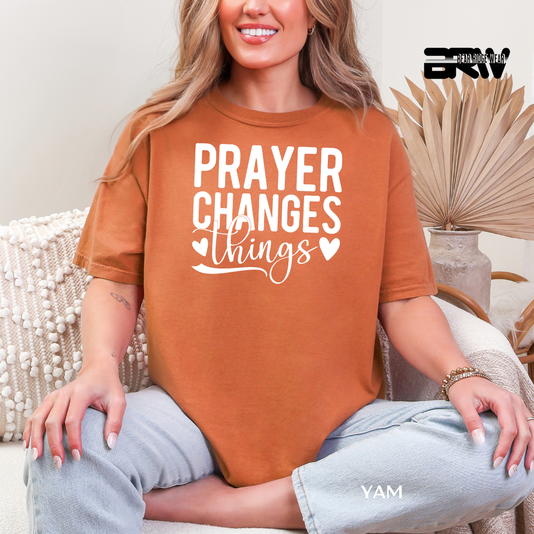 Woman wearing a brown t-shirt with 'PRAYER CHANGES things' text, sitting on a couch.