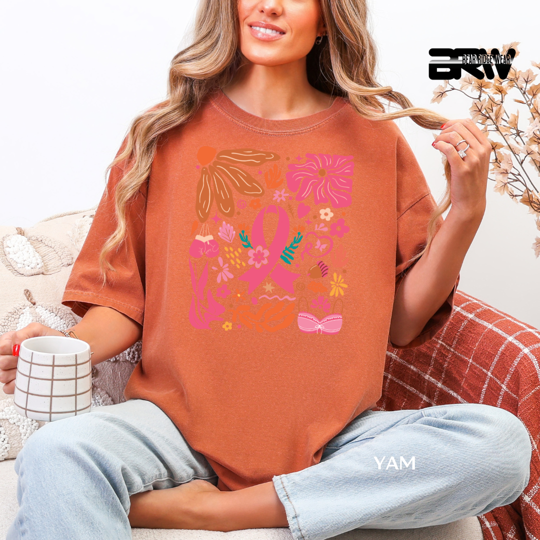 Woman wearing a brown t-shirt with colorful designs, sitting on a couch holding a mug. BREAST CANCER AWARENESS TEE 
