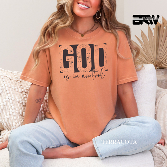 Woman wearing a terracotta t-shirt with 'GOD is in control' text, sitting on a couch.