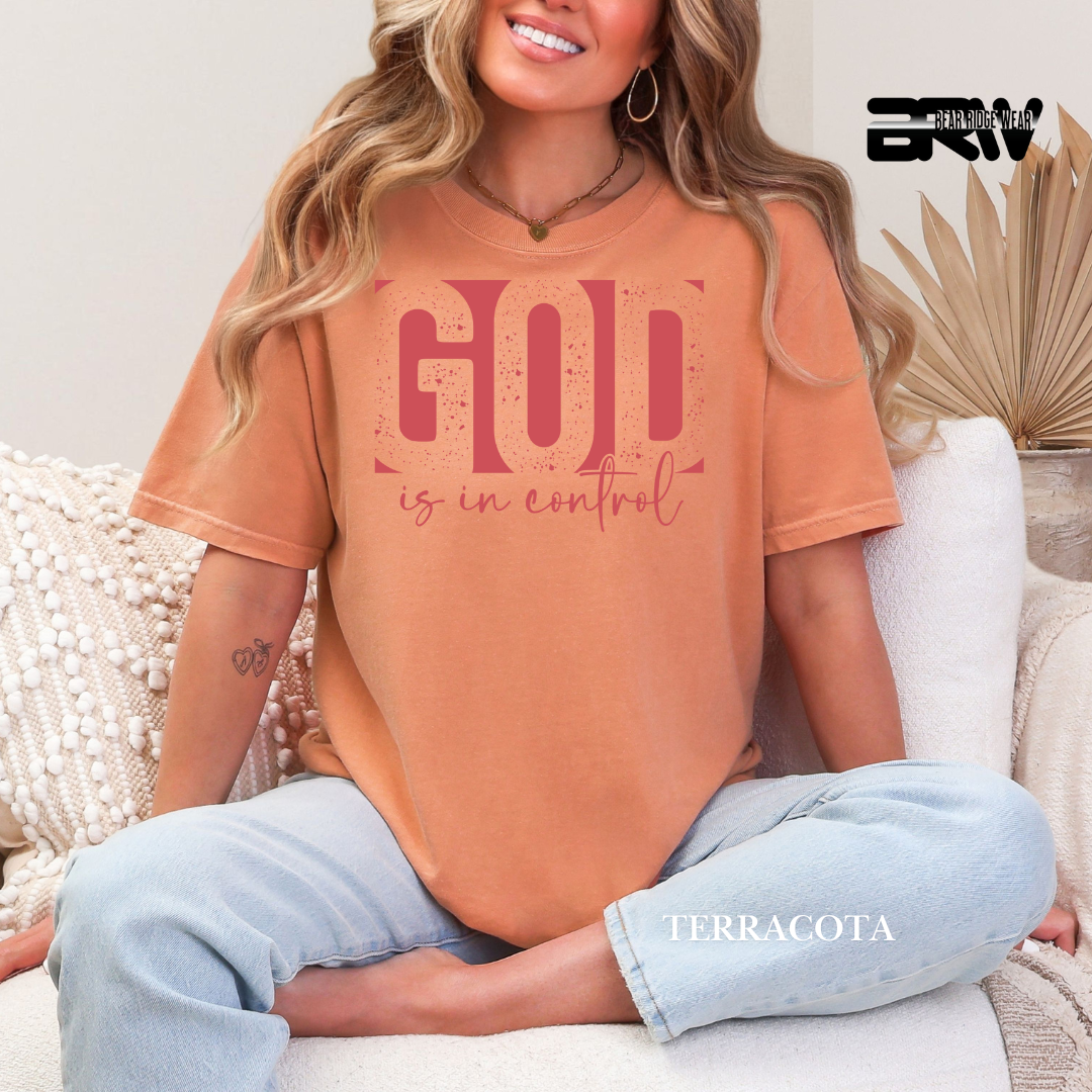 Woman wearing a terracotta t-shirt with 'God is in control' text, sitting on a couch.