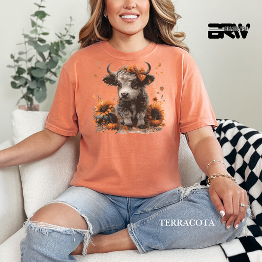 Woman wearing a terracotta-colored t-shirt with a cow and sunflower design, sitting on a couch.