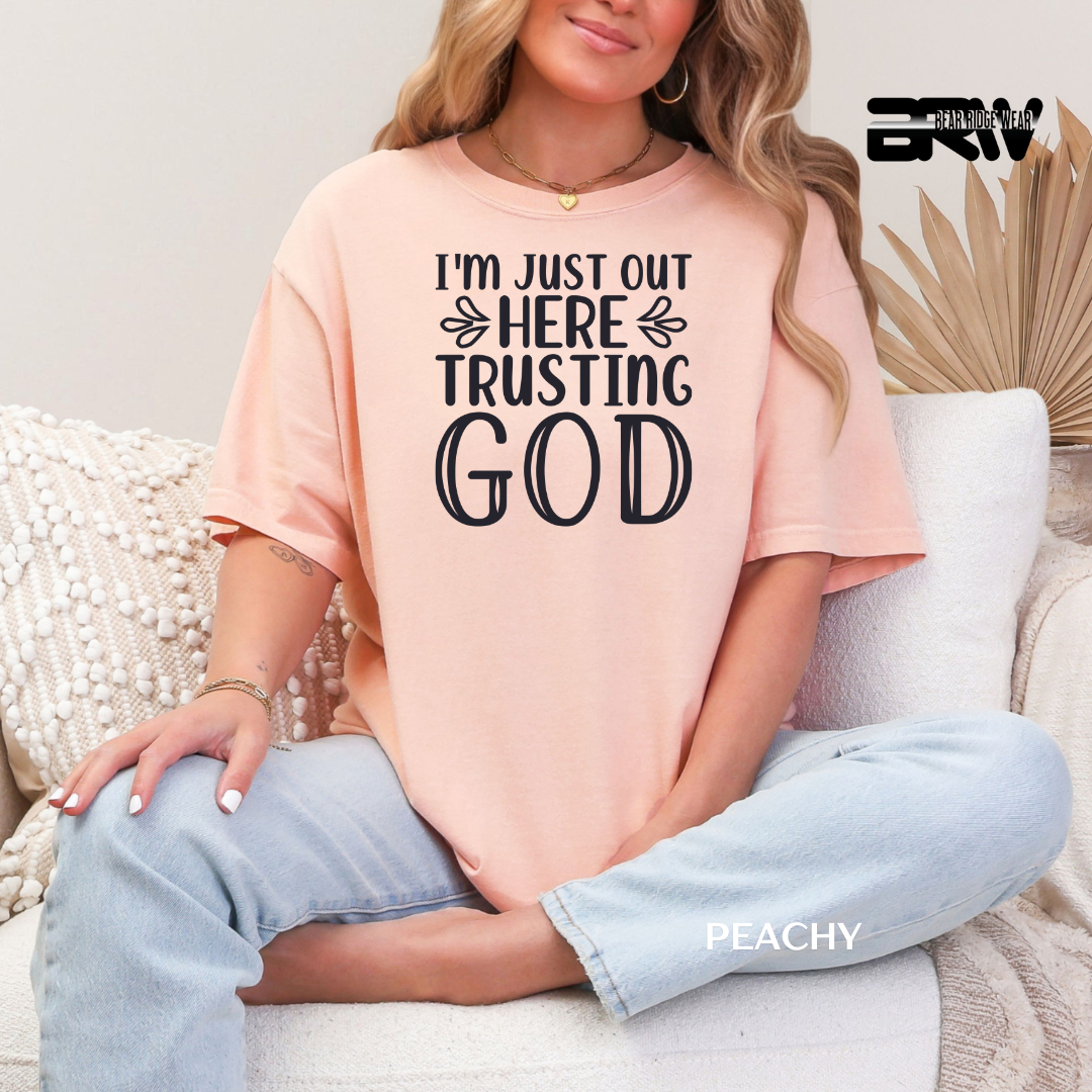Person wearing a peach t-shirt with text sitting on a couch. I'M JUST OUT HERE TRUSTING GOD FAITH TEE