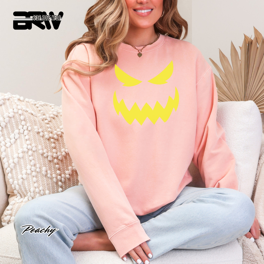 Person wearing a pink sweatshirt with a yellow graphic design, sitting on a couch. SCARY PUMPKIN FACE FALL TEE 