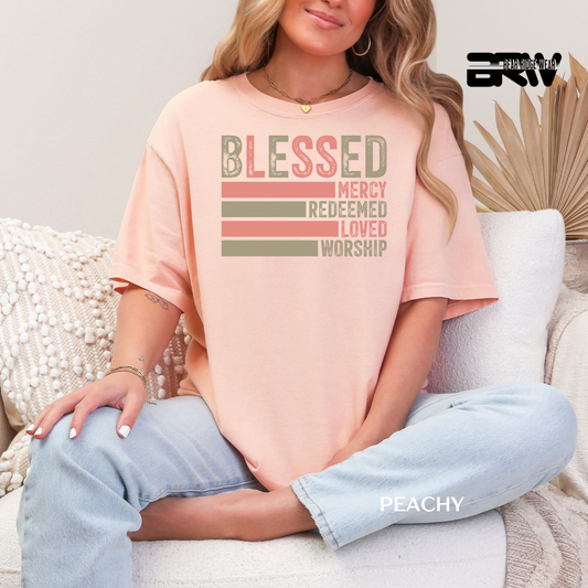 Person wearing a peach t-shirt with motivational text sitting on a couch. BLESSED, MERCY REDEEMED, LOVED AND WORSHIP FAITH TEE