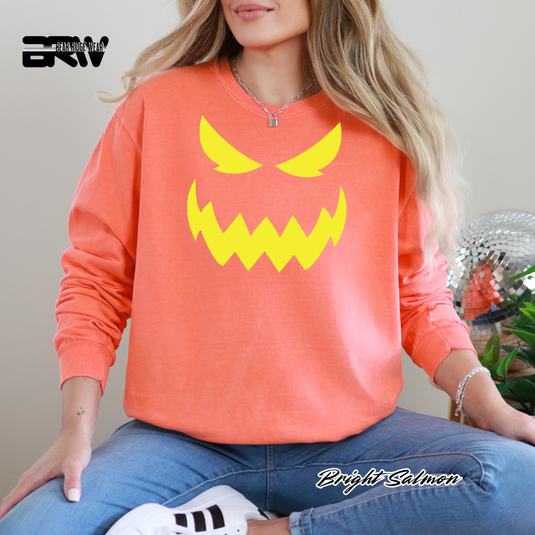 Person wearing a bright salmon-colored sweatshirt with a yellow graphic design, sitting on a chair. PUMPKIN FALL LONG SLEEVE TEE