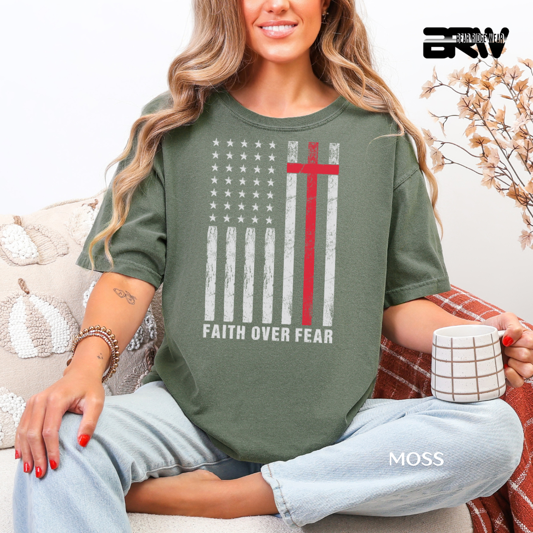Woman wearing a green t-shirt with a graphic design and text, sitting on a couch holding a mug. FAITH OVER FEAR WIH IUSA FLAG FAITH TEE