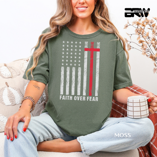 Woman wearing a green t-shirt with a graphic design and text, sitting on a couch holding a mug. FAITH OVER FEAR WIH IUSA FLAG FAITH TEE