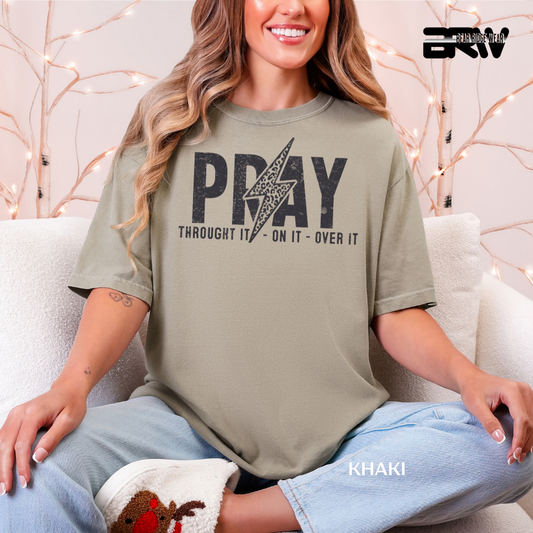 Woman wearing a khaki t-shirt with 'PRAY THROUGH IT, ON IT, OVER IT' text, sitting on a couch.