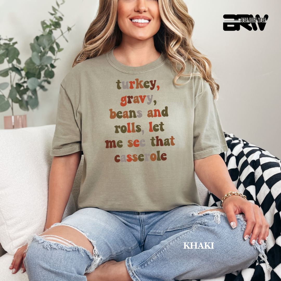 Woman wearing a khaki t-shirt with a humorous Thanksgiving message, sitting on a couch.
