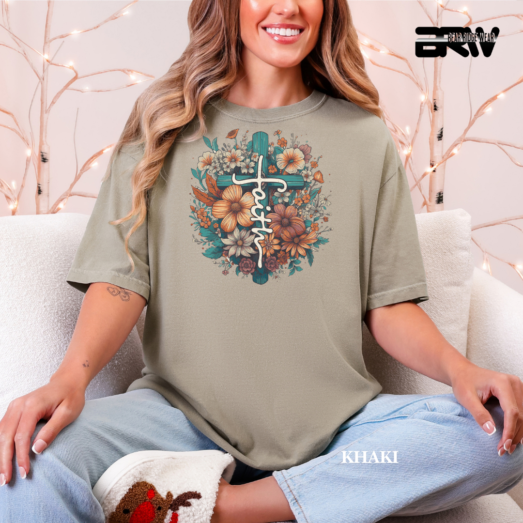Woman wearing a khaki t-shirt with a floral cross design and 'faith' text, sitting on a couch.